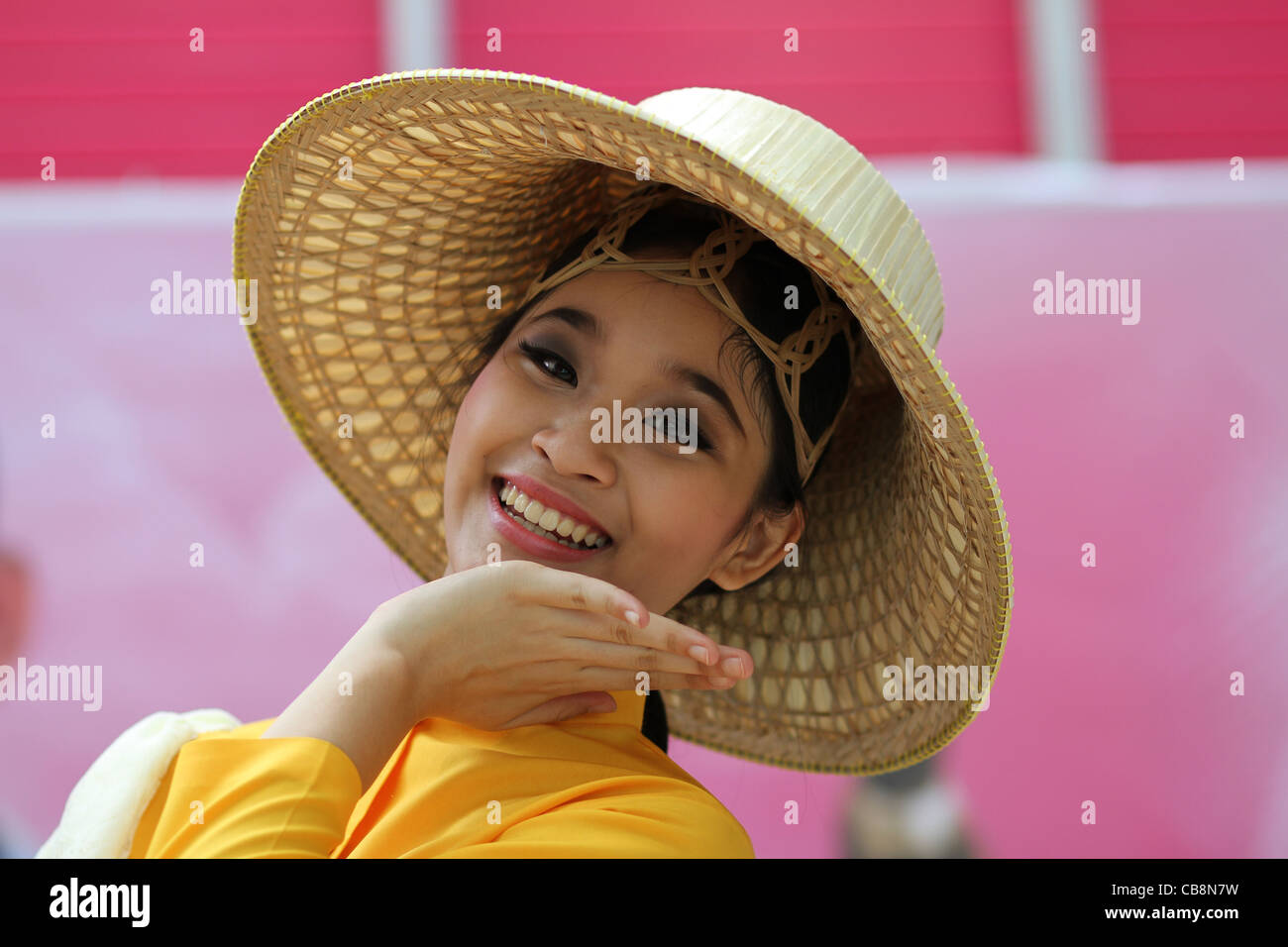 Ein thailändisches Mädchen in Tracht und trägt einen Korb Flechten Hut Gesten mit der Hand unter ihr Kinn, wie sie lächelt. Stockfoto