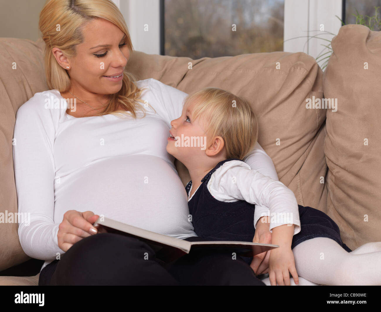 Junge schwangere Mutter und ihre drei Jahre alte Tochter, die zusammen ein Buch zu lesen, die zu Hause Stockfoto