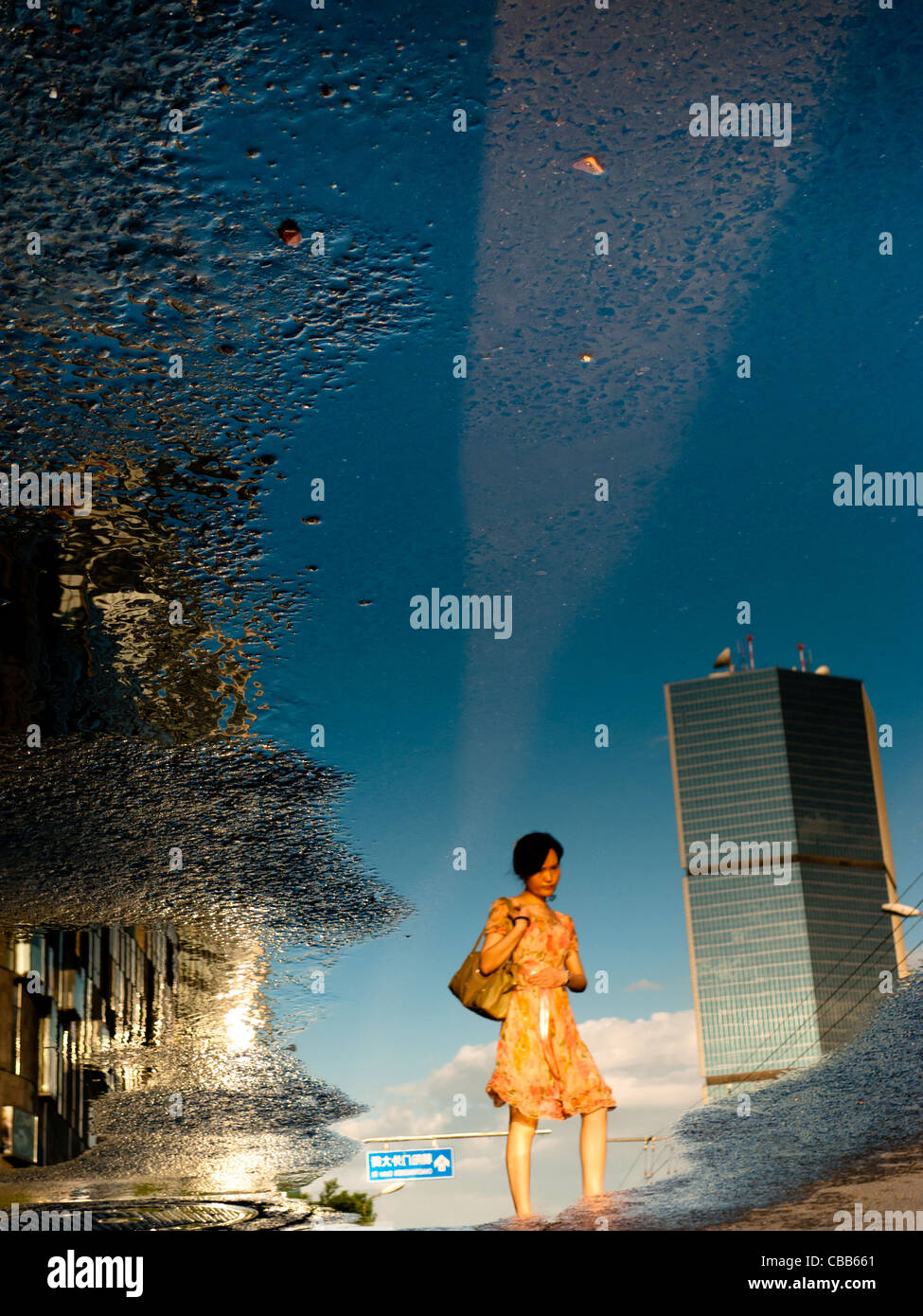 Spiegelbild in einer Pfütze eine nachdenkliche Frau. Im Hintergrund ein Wolkenkratzer vor blauem Himmel. Stockfoto