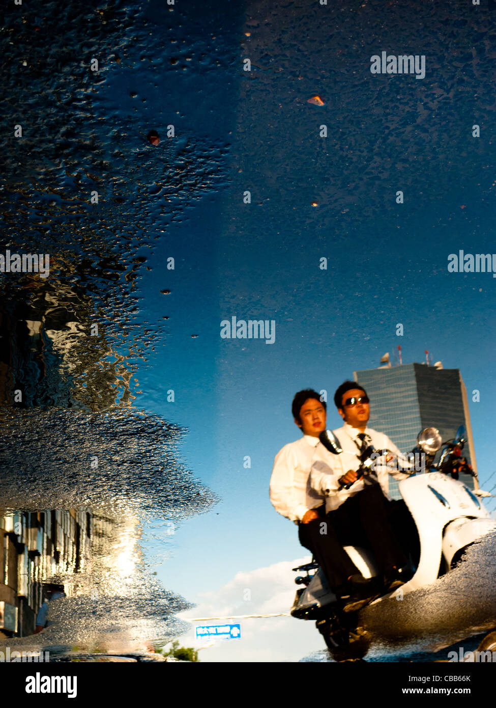 "California Dreaming". Spiegelbild in einer Pfütze der zwei Männer auf einem Roller mit, im Hintergrund, ein Wolkenkratzer vor blauem Himmel. Stockfoto
