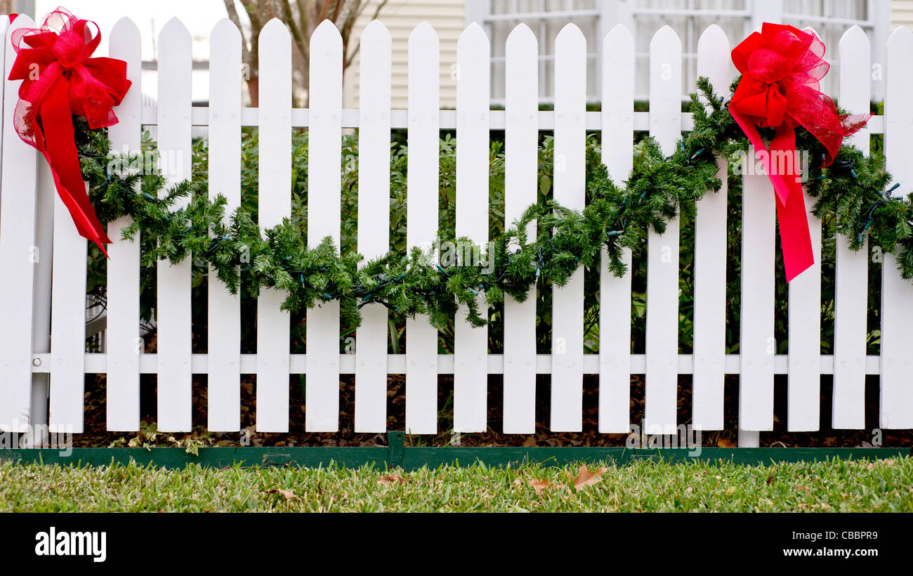 Garland und rote Schleifen hängen von einem weißen Lattenzaun für die Weihnachtszeit. Stockfoto