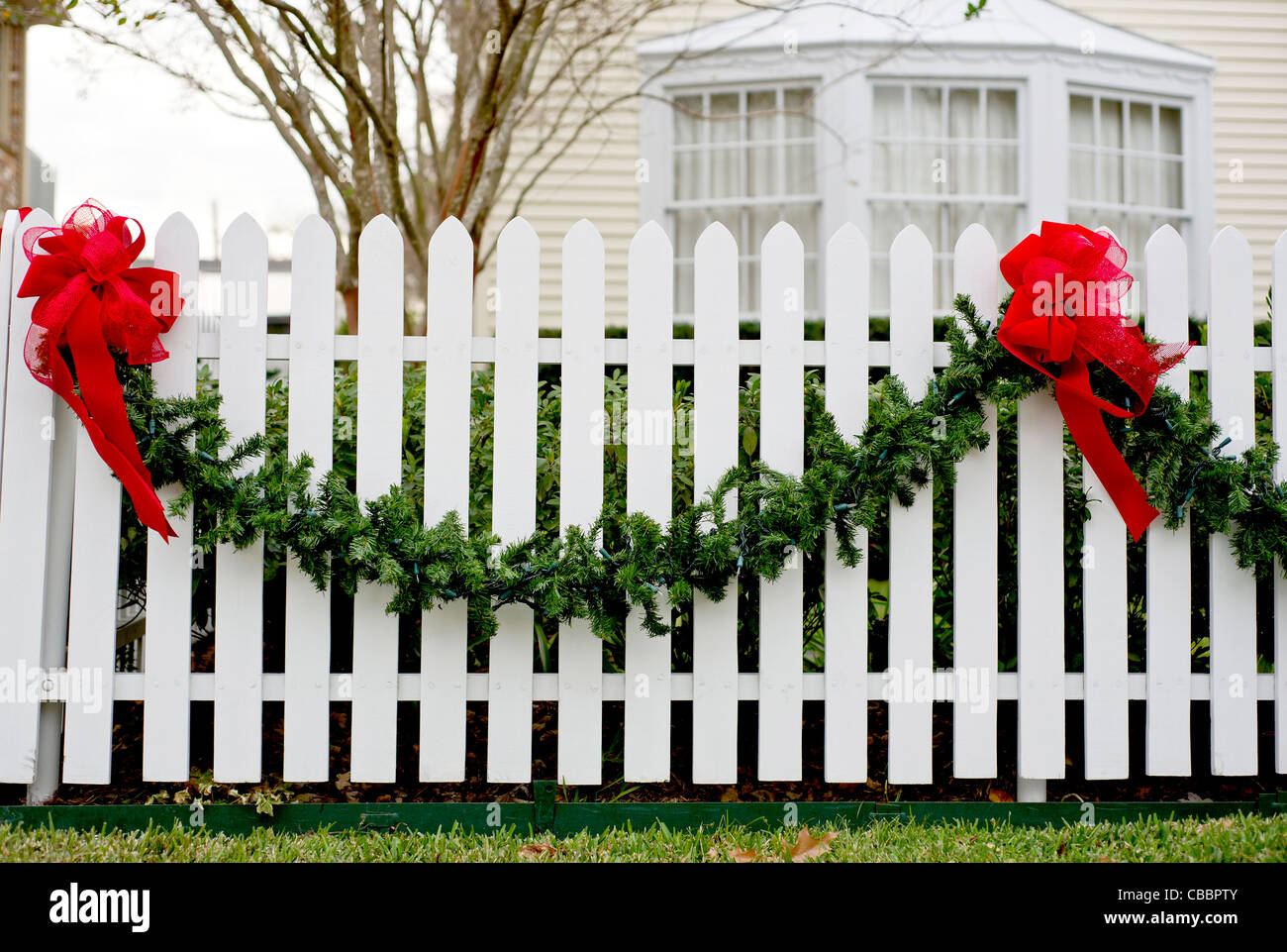 Garland und rote Schleifen hängen von einem weißen Lattenzaun für die Weihnachtszeit. Stockfoto