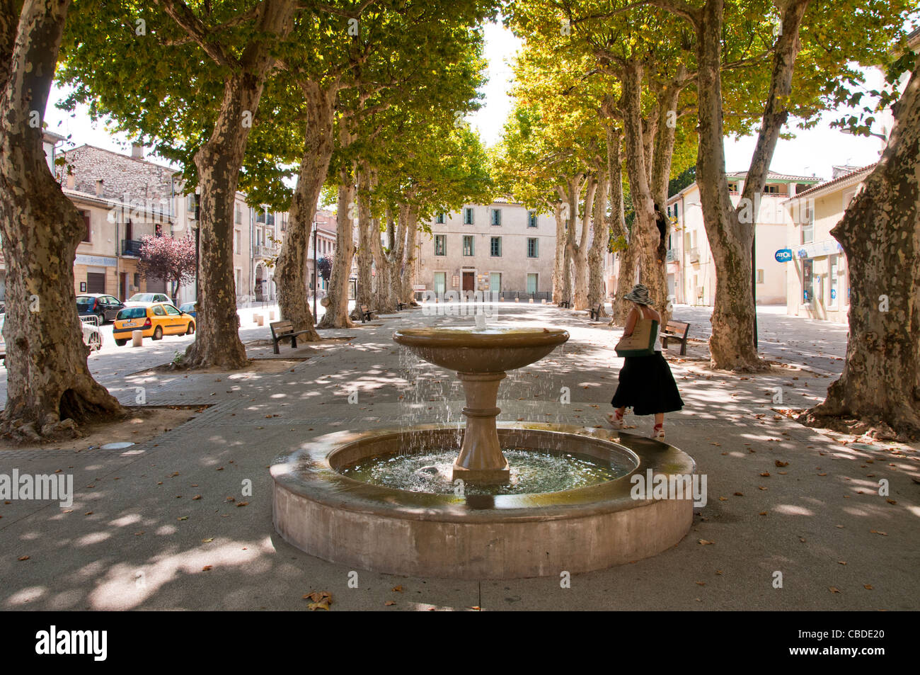 eine Frau, vorbei an einem Brunnen in einem Baum gesäumten Platz in Gignac Gignac Hérault Languedoc-Roussillon Frankreich Stockfoto