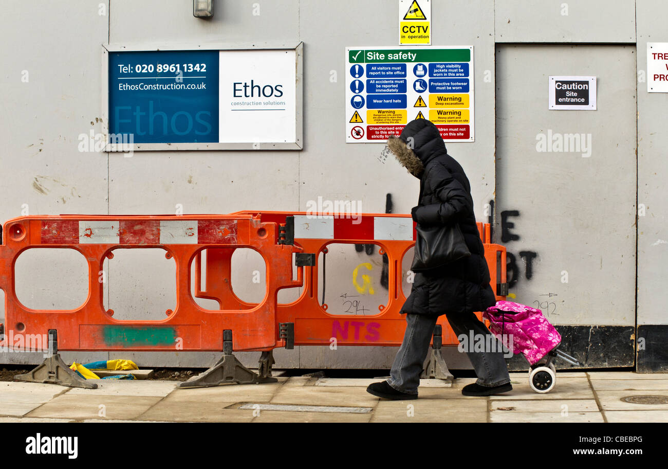 Kapuzengestalt zu Fuß vor einer Baustelle mit Sicherheitshinweisen, ziehen eine rosa Einkaufswagen Stockfoto