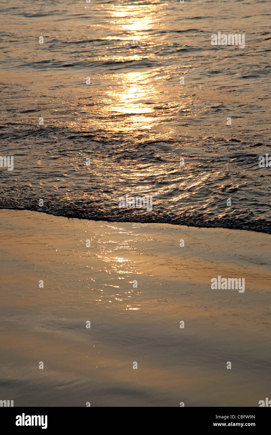 Wellen brechen sich an einem ruhigen Sandstrand mit der Reflexion der Sonne auf das Wasser des Meeres bei Sonnenuntergang am Patong Beach in Phuket Stockfoto