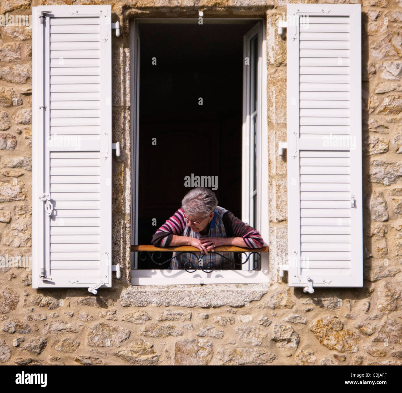 Alte Frau schaut aus einem Fensterläden Fenstertür in Frankreich Stockfoto
