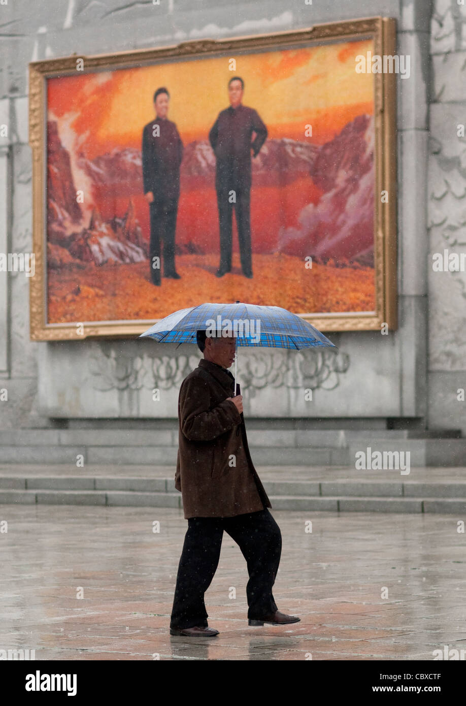 MANN MIT EINEM REGENSCHIRM VOR EIN PROPAGANDAPLAKAT, PYONGYANG, NORDKOREA Stockfoto