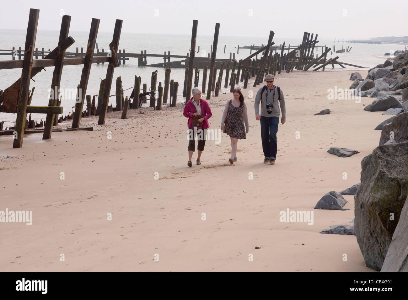 Happisburgh Küste, North Norfolk, East Anglia. Drei Menschen zu Fuß zwischen nicht Holz und Metall Leistungsschalter, Buhnen; importierte Felsen rechts Stockfoto