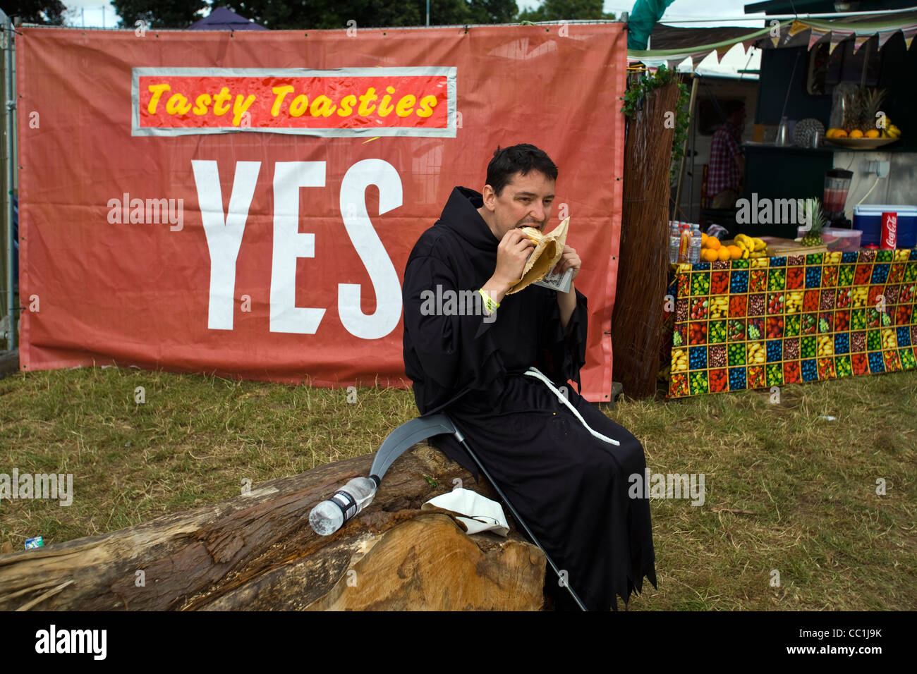 Ein Mann verkleidet als der Sensenmann auf einem Baumstamm auf dem Standon Calling Festival in Hertfordshire, Großbritannien einen Sandwich isst. Stockfoto
