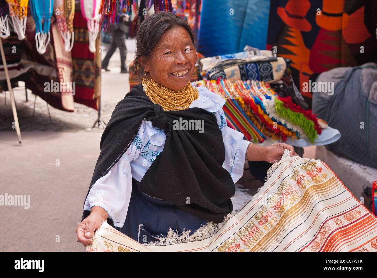 Eine indische Frau lächelt und zeigt einen gewebten Teppich zum Verkauf auf dem Markt in Otavalo, Ecuador. Stockfoto