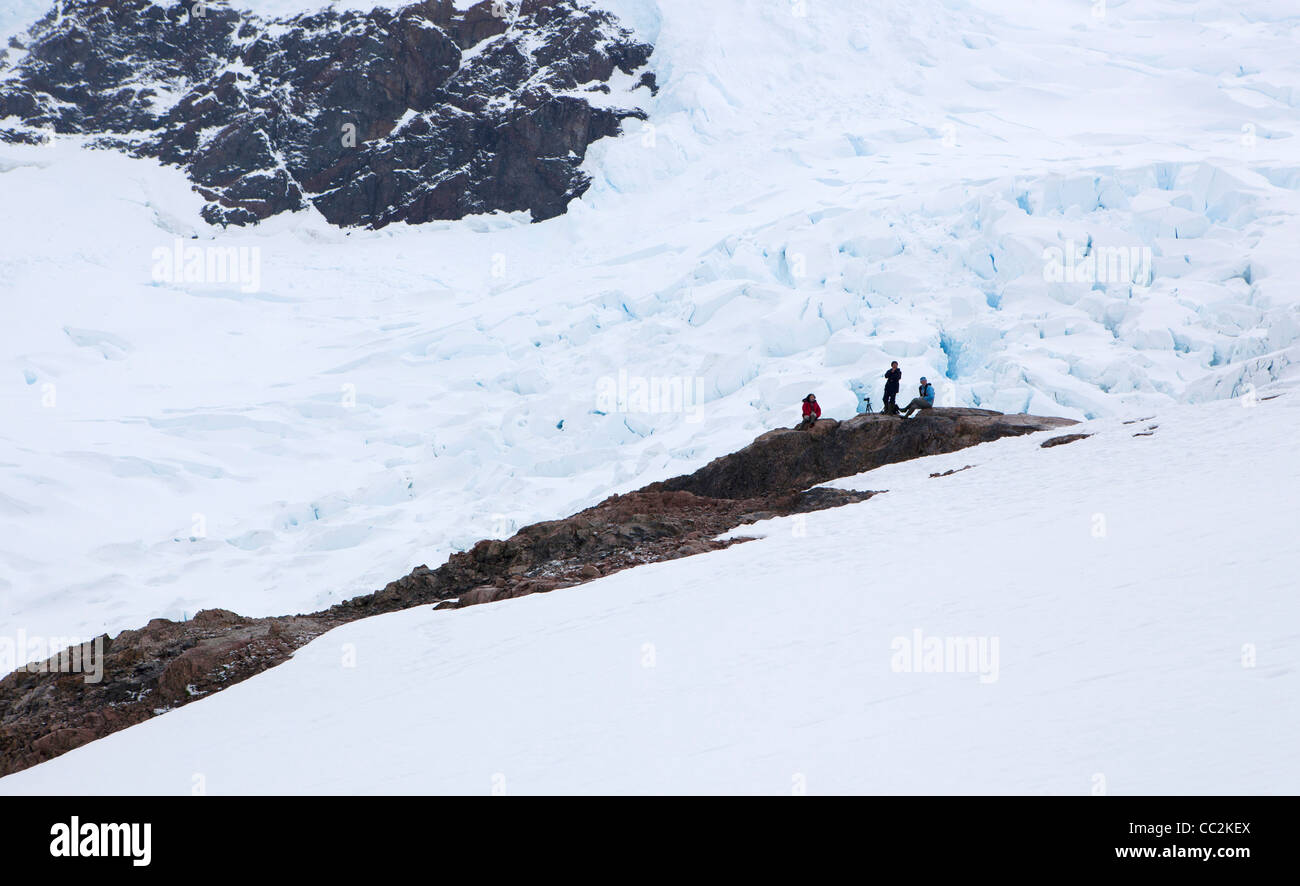 Touristen sitzen auf einem Felsvorsprung Look neben einem Gletscher über Neko Harbour, Antarktis Stockfoto