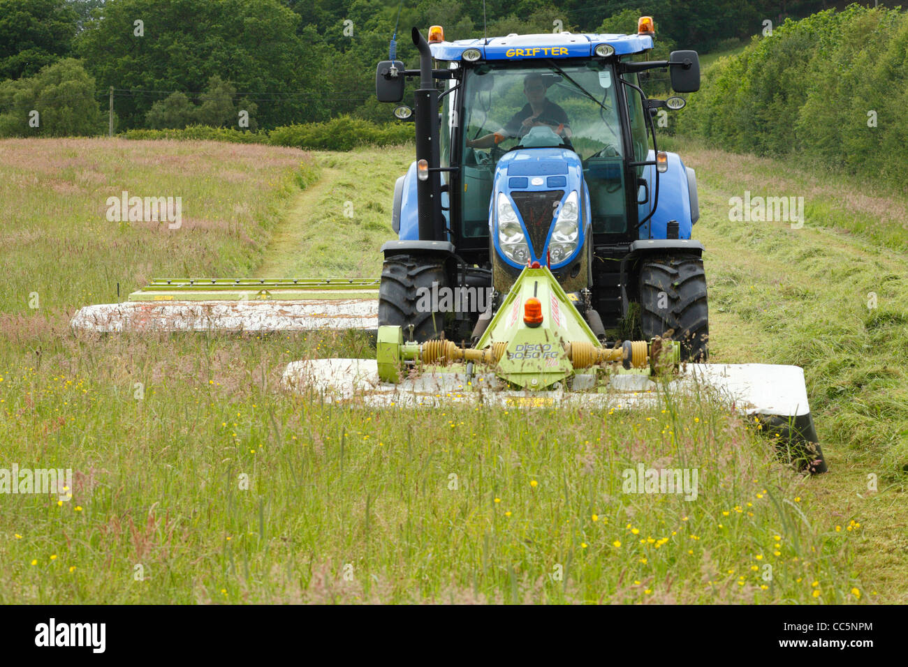 Auftragnehmer mit New Holland T6080 Traktor und vorne und hinten montiert Claas Mäher mäht Rasen für die Silage. Auf einem Bio-Bauernhof. Stockfoto