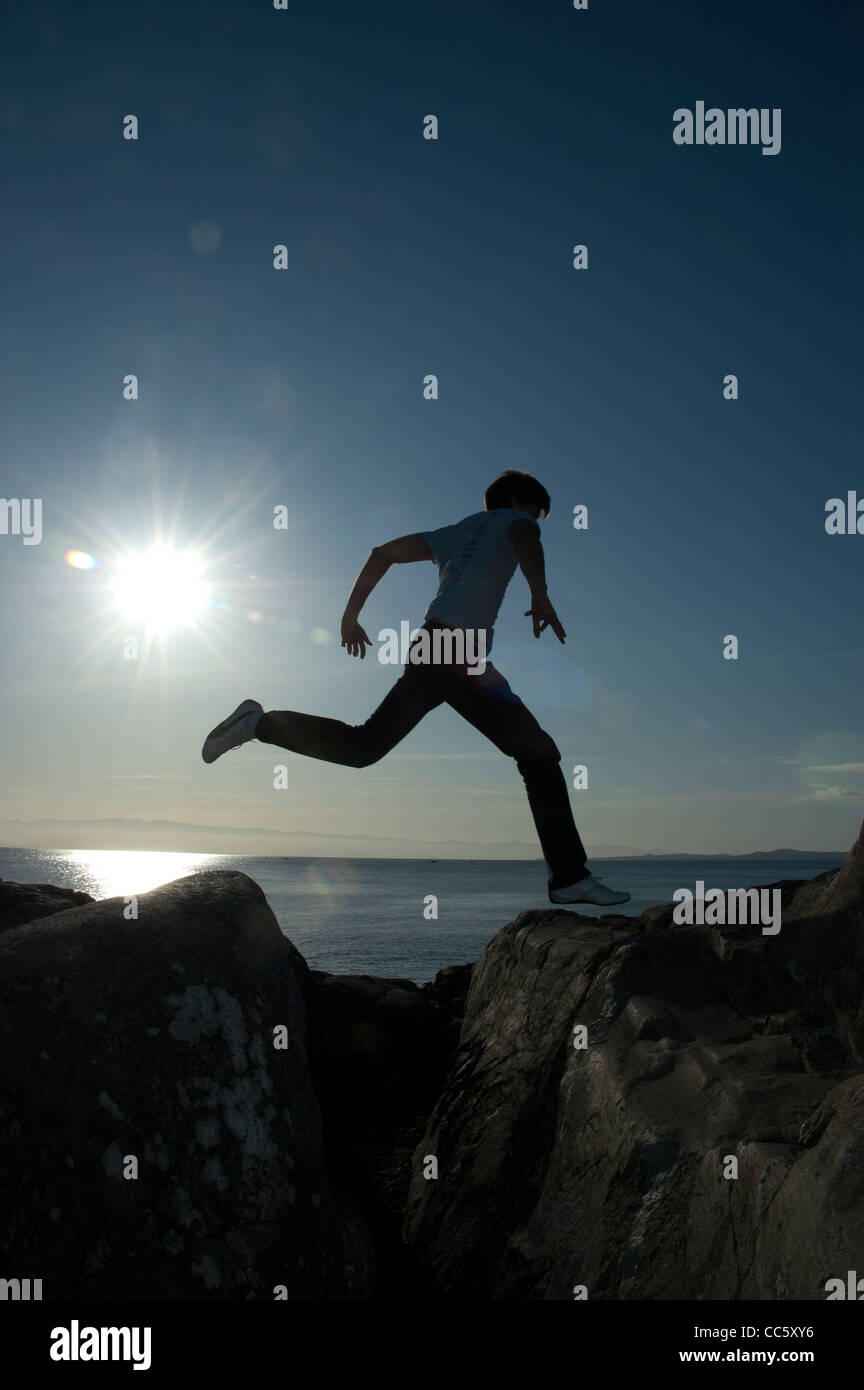 Eine niedrige Sonne Hintergrundbeleuchtung ein junger Mann über eine Lücke zu springen oder Harpunen in Rock, Beacon Hill Park, Victoria, Britisch-Kolumbien, Kanada. Stockfoto