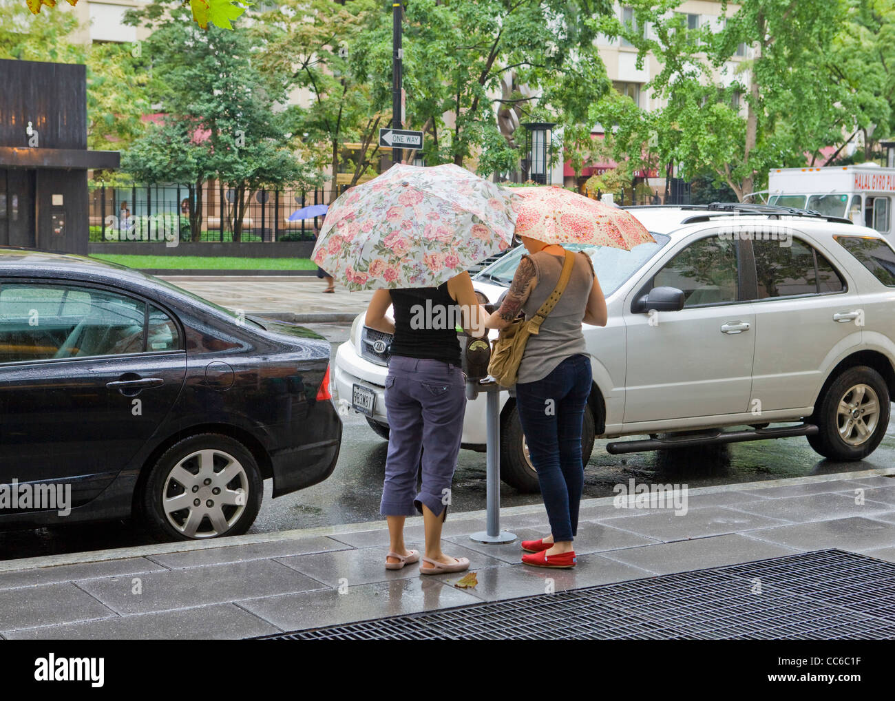 Zwei Frauen, die Fütterung einer Parkuhr halten Sonnenschirme an einem regnerischen Tag Stockfoto