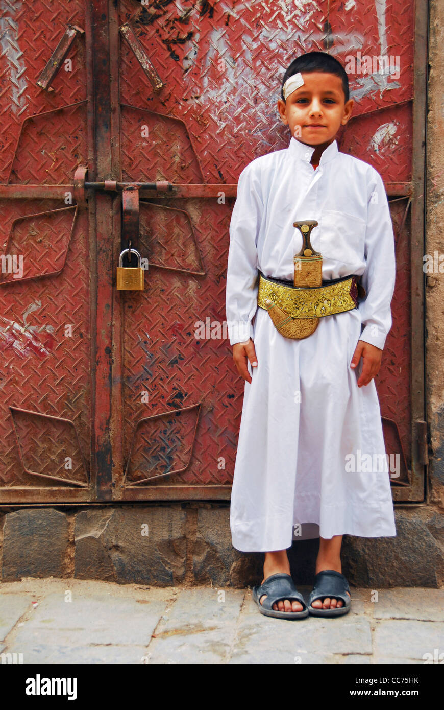 Jemen, Sanaa, Portrait eines wenig traditionell gekleideten Jungen stand vor verschlossenen metallischen Tür Stockfoto