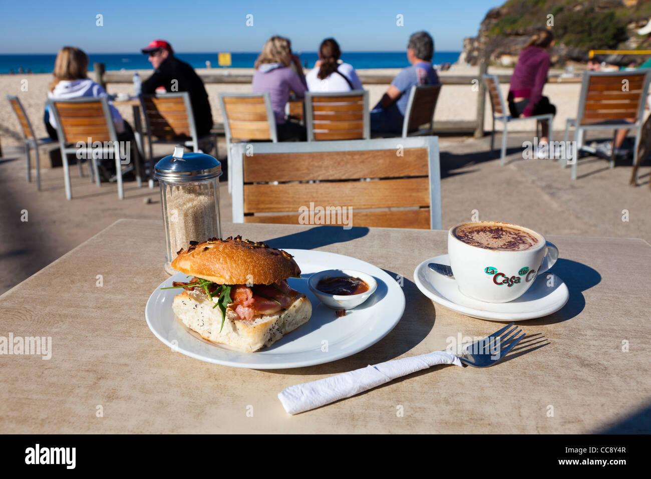 Frühstück in der Nähe: Tamarama Beach Sydney mit Kaffee und Sandwich. Draußen am Tisch mit Leuten im Hintergrund Stockfoto