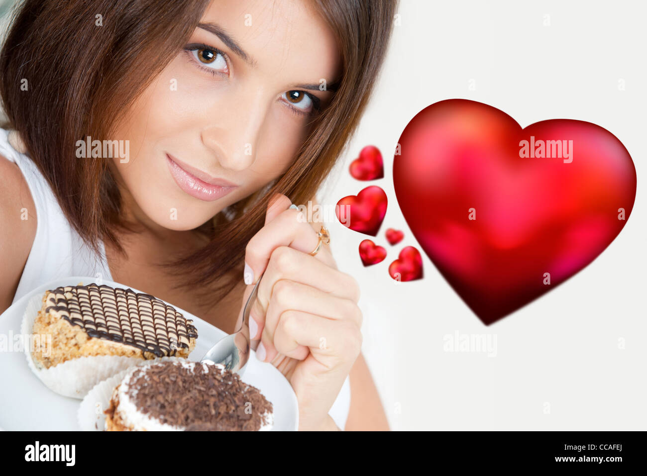 Lächelnde Brünette Frau Essen ein Stück Kuchen im Wohnzimmer in ihrer Wohnung. Rotes Herz Grafiksymbole 3d fliegen in ihrer Nähe. Stockfoto
