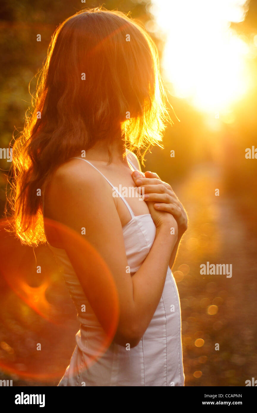 Schöne junge Mädchen mit langen Haaren und weißen Kleid mit Blick auf das goldene Licht der untergehenden Sonne mit Händen auf ihr Herz Stockfoto