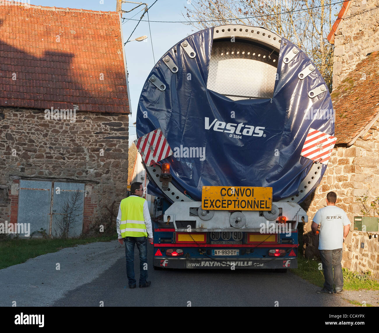 Außergewöhnlicher Transport eines Pylonelements der Windturbine. Zwei Fahrer überwachen und führen den Anhänger, um diesen sehr schmalen Durchgang zwischen zwei Häusern zu gewährleisten Stockfoto Außergewöhnlicher Transport eines Pylonelements der Windturbine. Zwei Fahrer überwachen und führen den Anhänger, um diesen sehr schmalen Durchgang zwischen zwei Häusern zu gewährleisten Stockfoto
