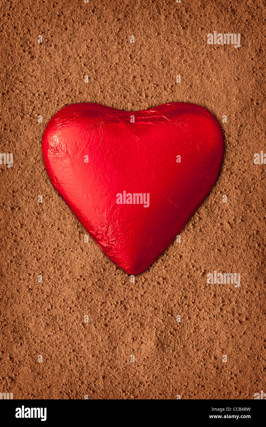 Rote Folie eingewickelt Schokoladenherz auf einem strukturierten Hintergrund von Coco Pulver gemacht Stockfoto