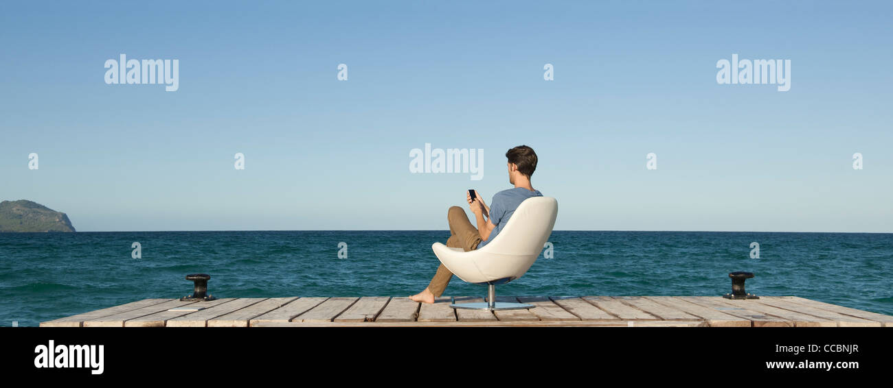 Mann Siitng im Stuhl auf dock mit Handy, Rückansicht Stockfoto