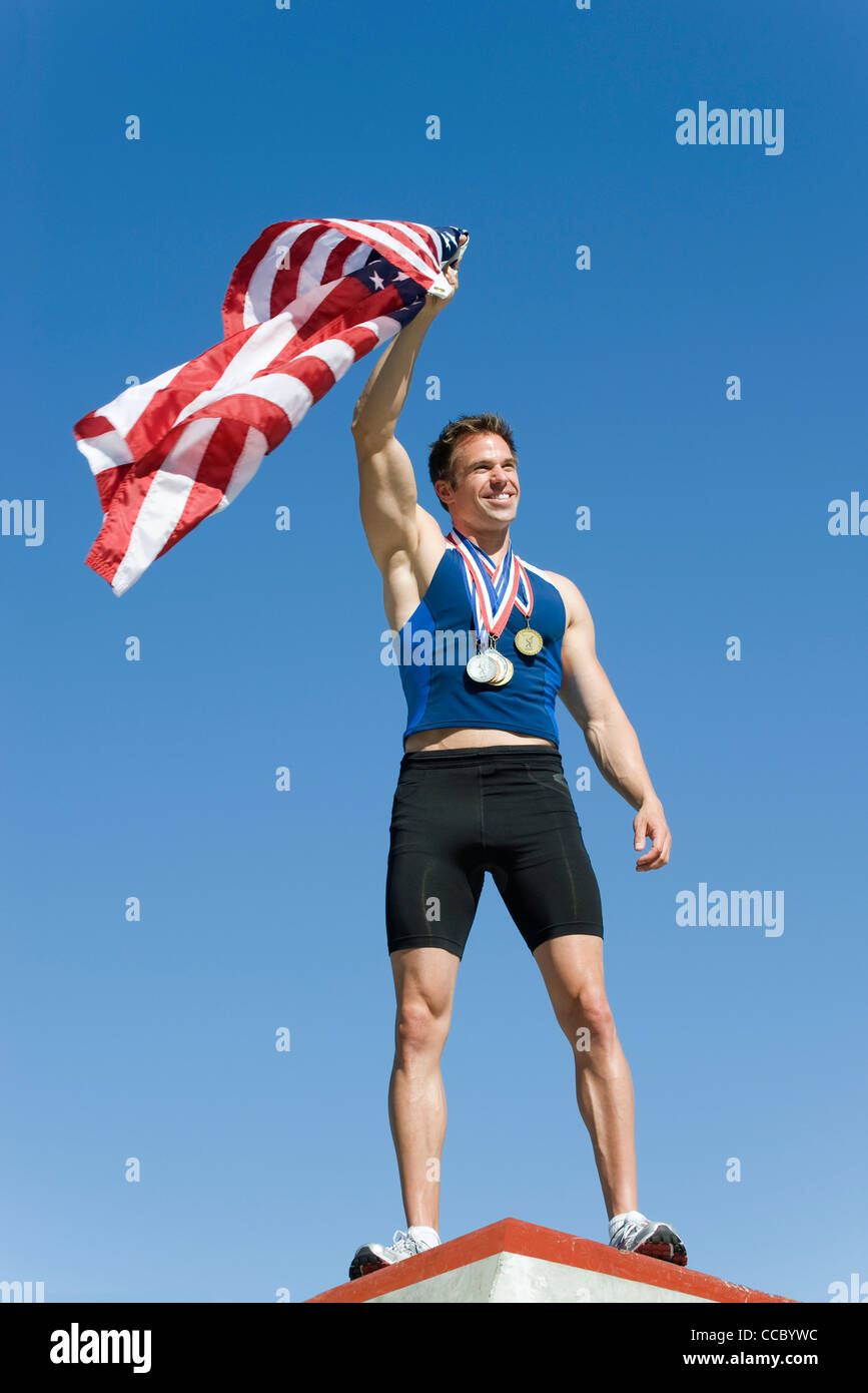 Männlicher Athlet auf Siegertreppchen, amerikanische Fahne hochhalten Stockfoto