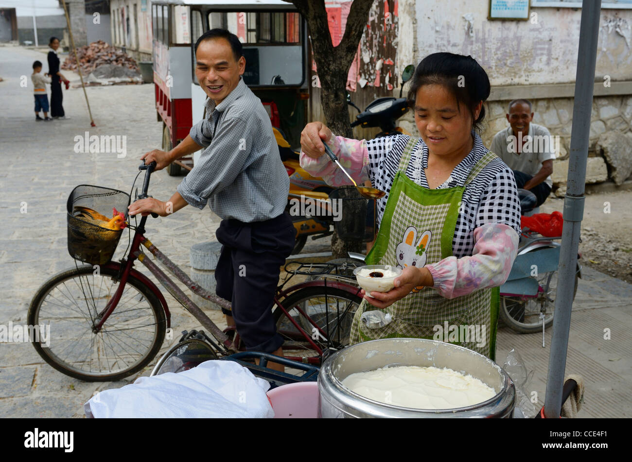 Straßenverkäufer Ladelling Sirup auf frischem Quark Douhua mit Nachbarn auf Fahrrad mit lebenden Hühnern in Fuli China Stockfoto