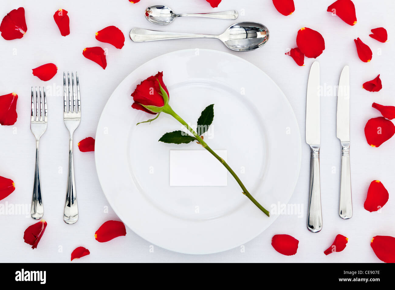 Foto von einer Tabelle Gedeck mit einer roten rose und leere Karte auf dem Teller plus Rosenblätter auf der Tischdecke Stockfoto