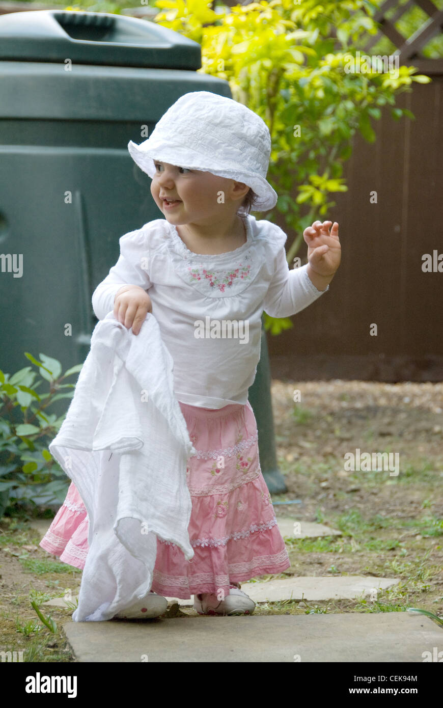 Ein weibliches Baby, tragen eine weiße Sommerhut, weißen Top und rosa Rock, stehen im Garten, hält ein Musselin-Quadrat, lächelnd, Stockfoto