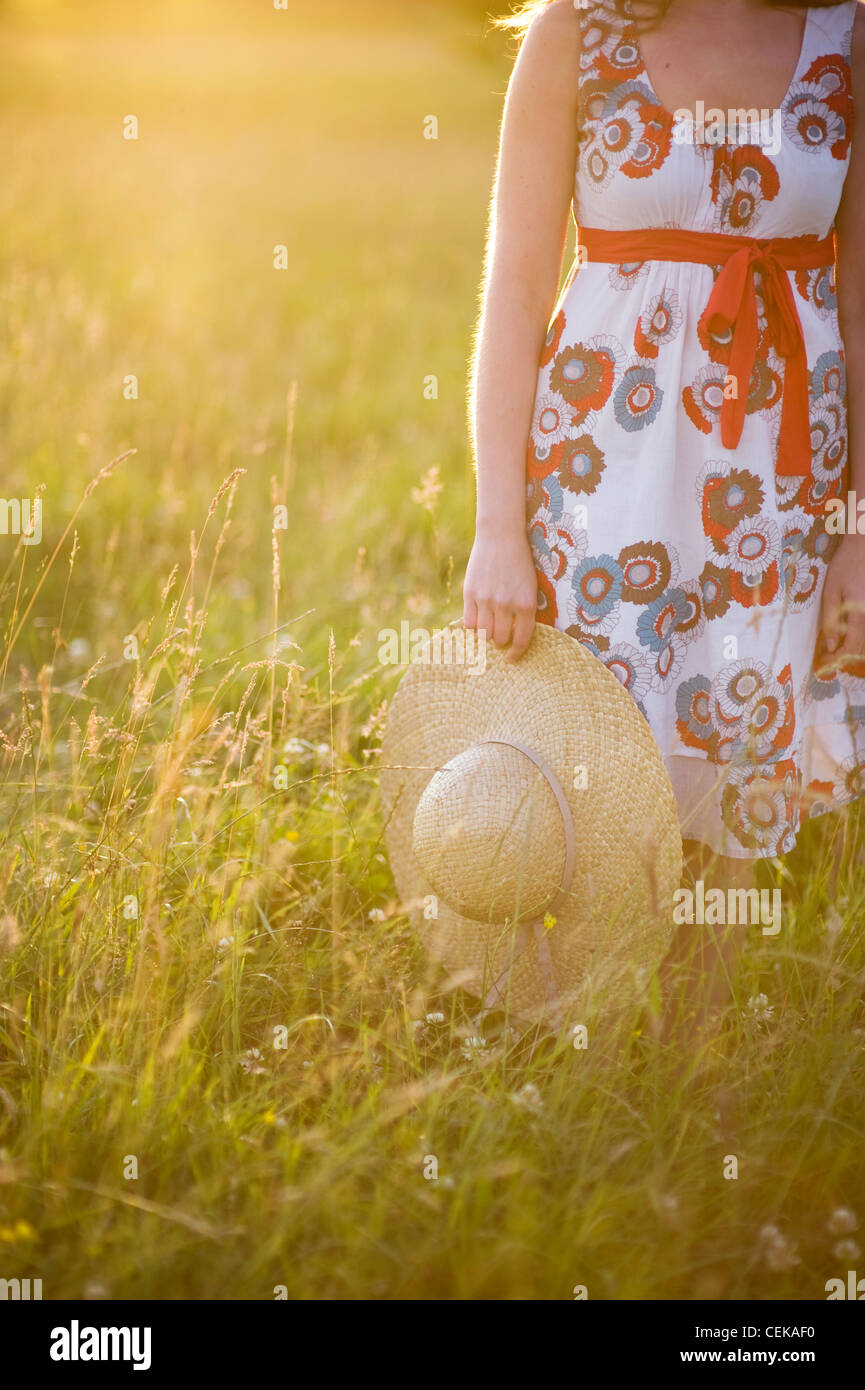 Frau stehend in einer Sommerwiese trägt eine Kleid und hält einen Stroh Sonnenhut Stockfoto