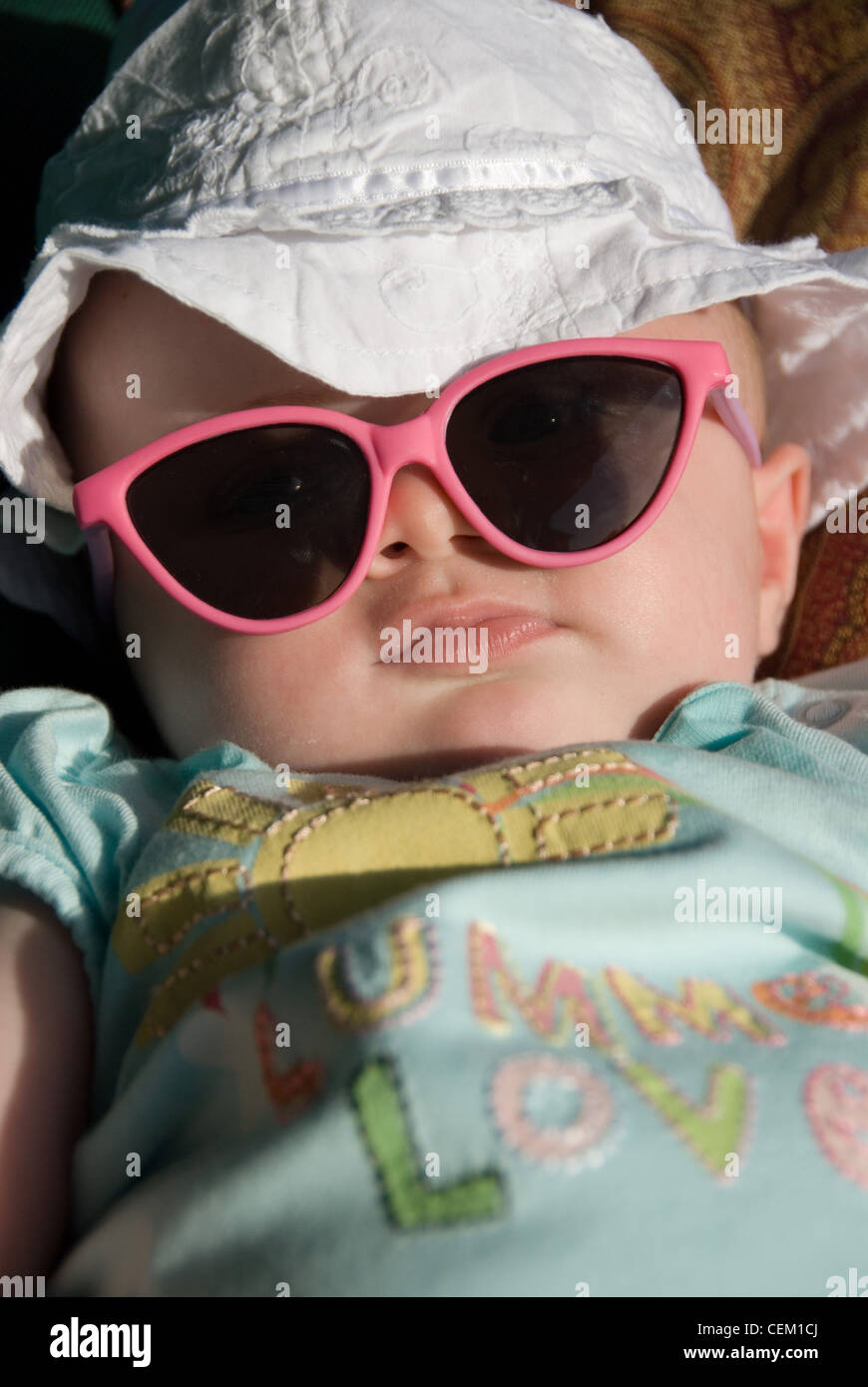 Ein weibliches Baby gekleidet in weißen Sonnenhut, ein blaues Muster-t-Shirt und ein großes Paar rosa umrandeten sunglasss Stockfoto