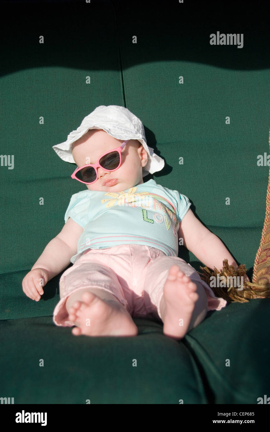 Ein weibliches Baby gekleidet in weißen Sonnenhut, ein blaues Muster-t-Shirt, rosa Hose und ein großes Paar rosa umrandeten sunglasss Stockfoto