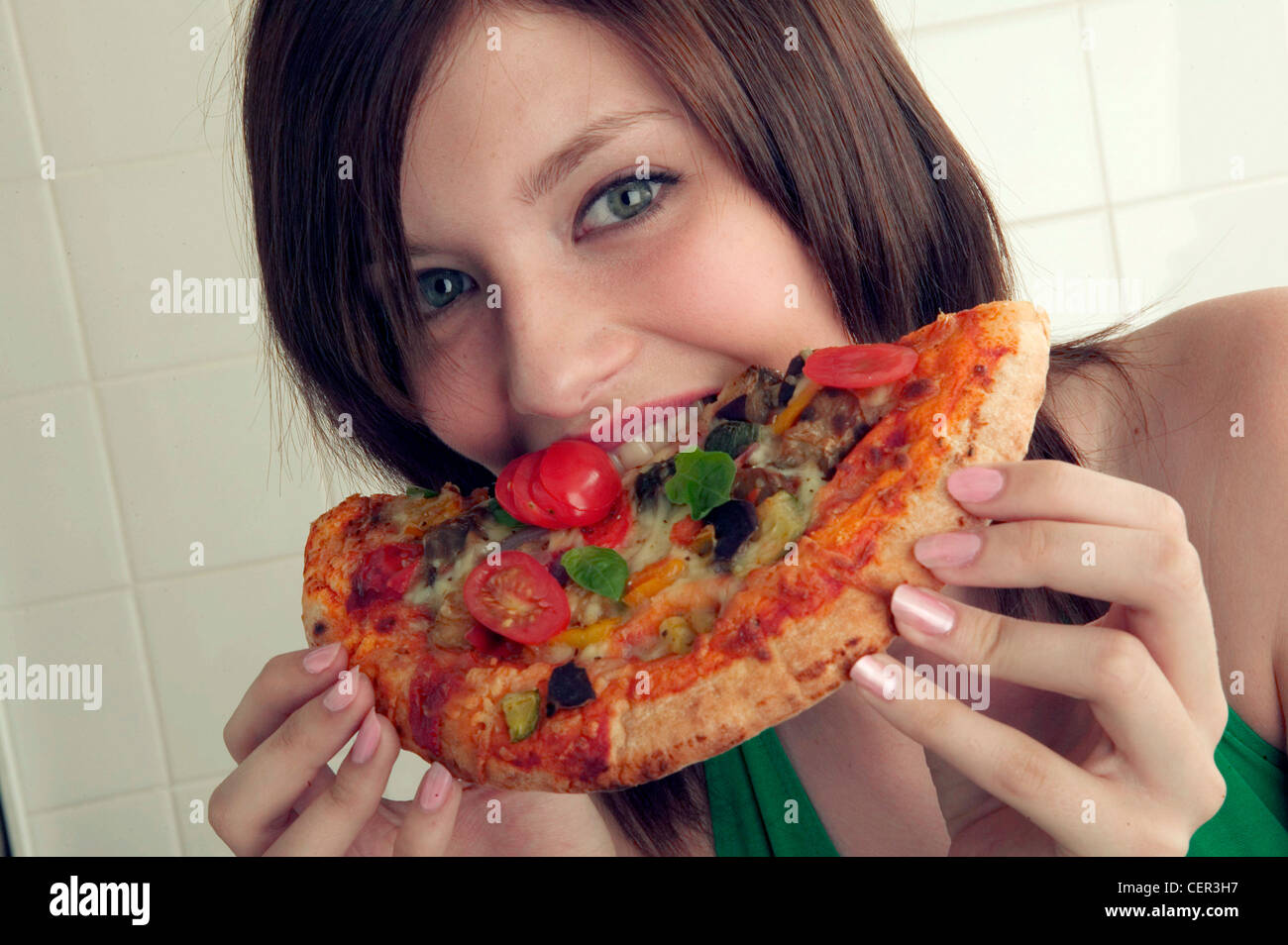 Frau mit schulterlangen Brünette Haaren, auf Kamera zum Anbeißen aus eine halbe Pizza, weiß geflieste Wand im Hintergrund Stockfoto