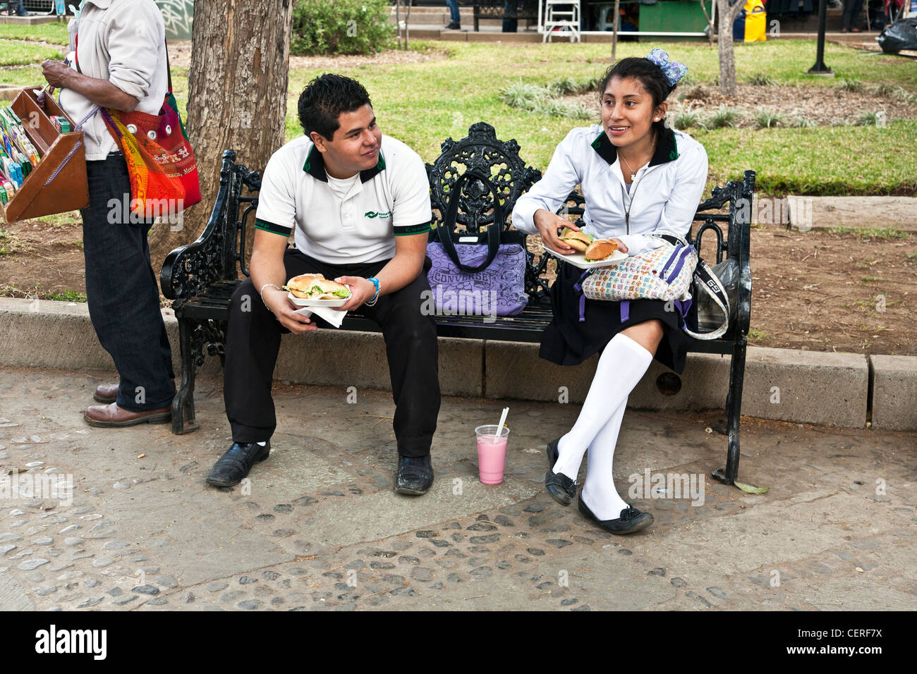 junge & Mädchen-technical High School Schüler in Schuluniform Mittagessen auf einer viktorianischen Schmiedeeisen Bank in Llano Park Oaxaca Stockfoto