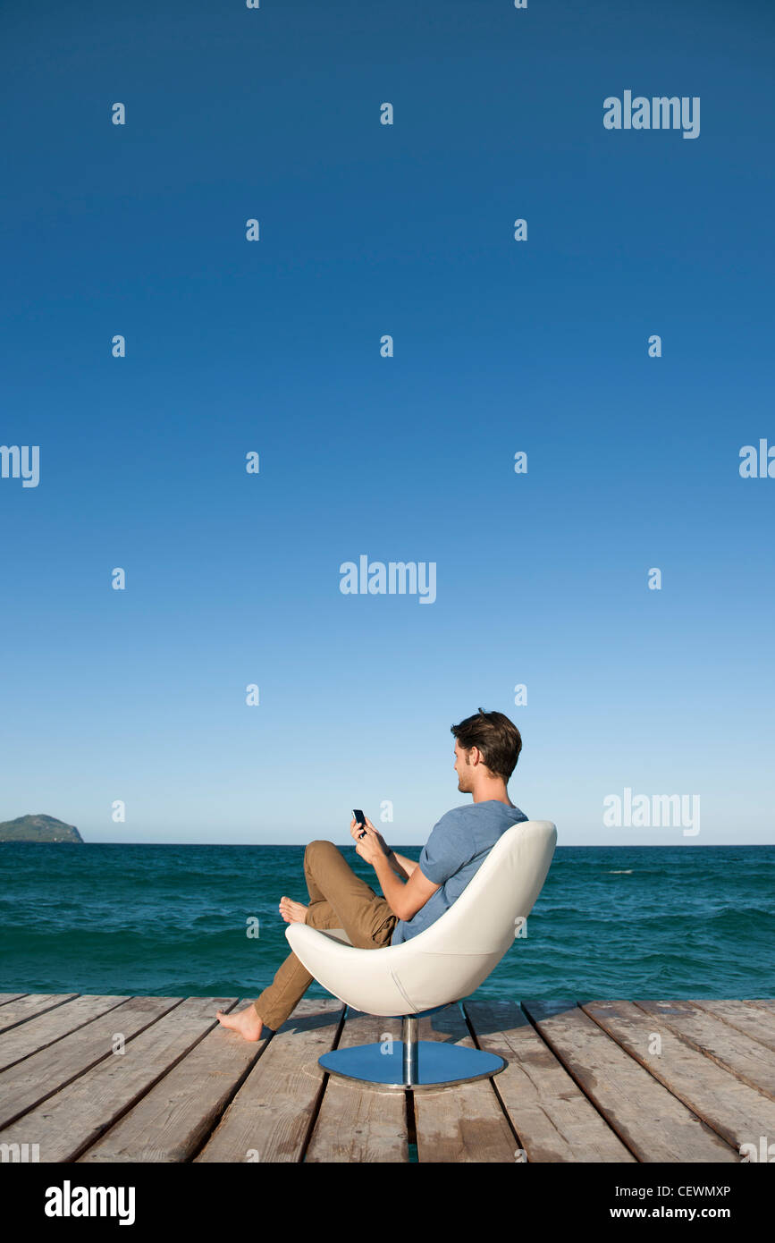 Junger Mann sitzt im Sessel See mit Handy, Seitenansicht Stockfoto