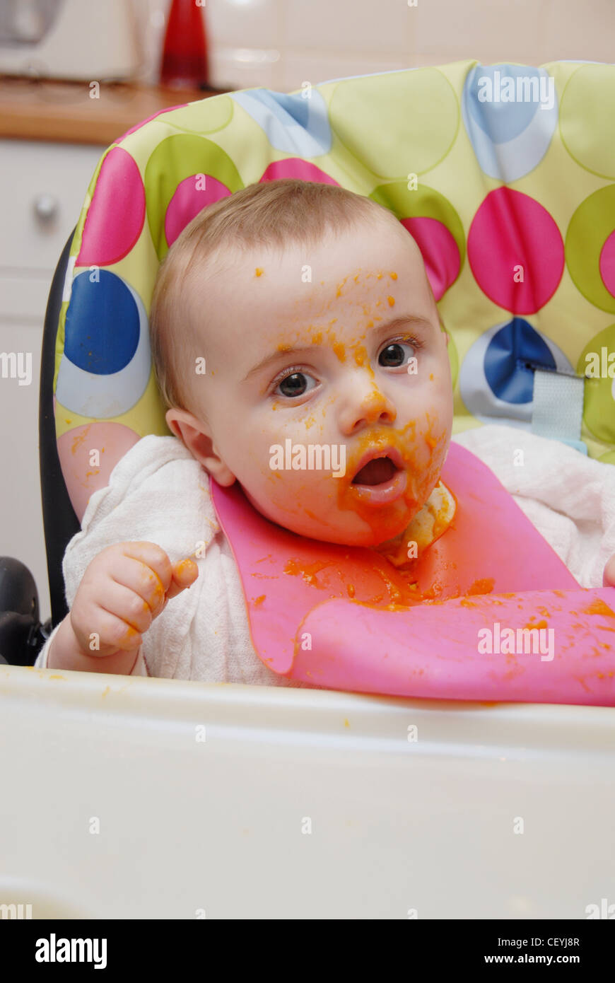 Ein weibliches Baby blondes Haar, im Hochstuhl sitzen, tragen eine quadratische Musselin und rosa Kunststoff-Lätzchen, püriert Essen Orange gefärbt Stockfoto