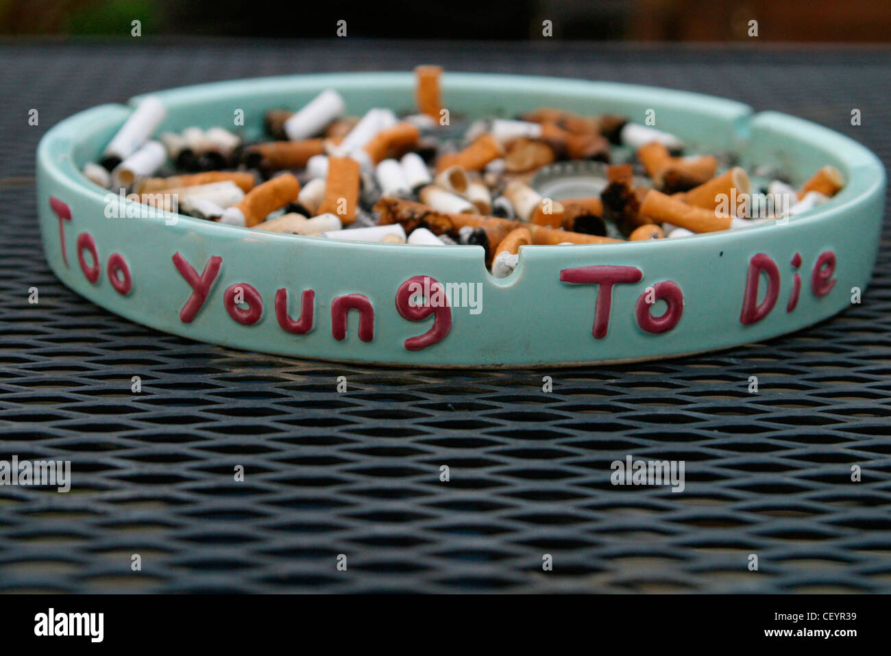 Hell blau Aschenbecher voller gerodet, Zigarettenkippen die Worte zu jung zum sterben, geschrieben auf der Seite in Rosa auf einem Draht-Tisch Stockfoto