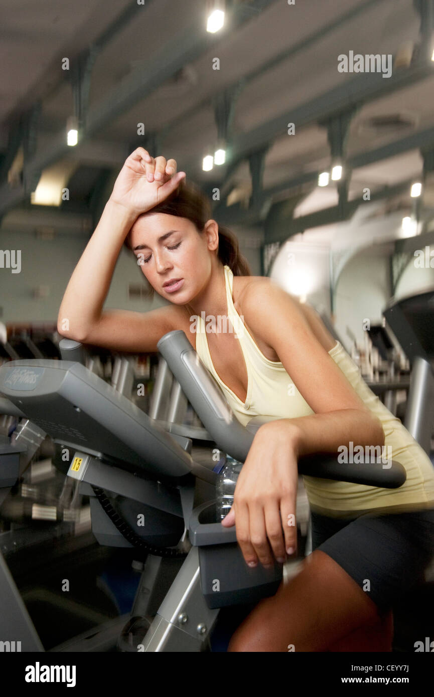 Weibliche brünette Haare gebunden zu Pferdeschwanz tragen eine gelbe Neckholder Top und graue Shorts, sitzen auf Heimtrainer, schiefe Stockfoto