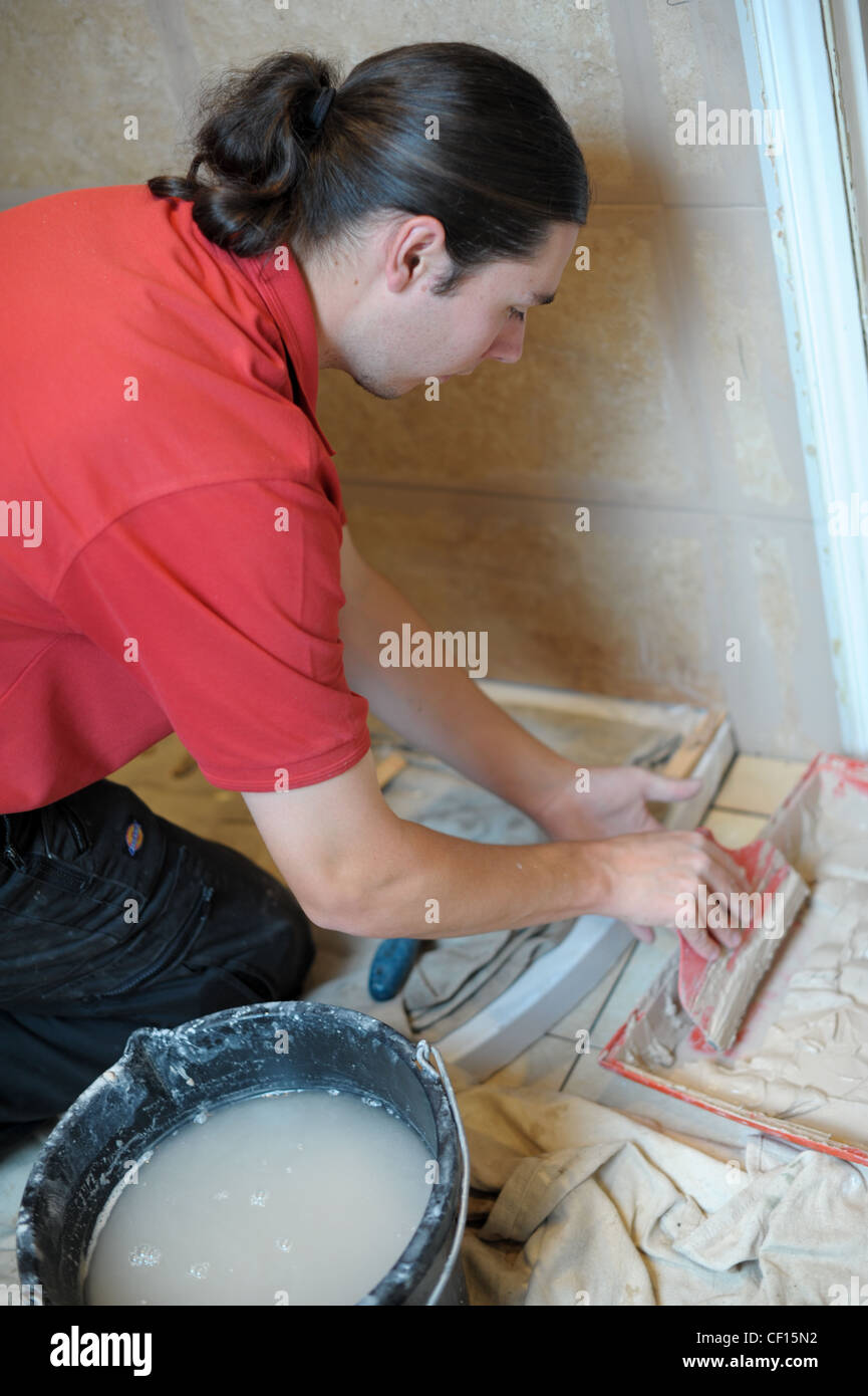 Mann kniet am Arbeitsplatz Verfugen der Fliesen in einem Badezimmer mit einem Eimer Wasser und ein Tablett mit Fugen auf dem Boden Stockfoto