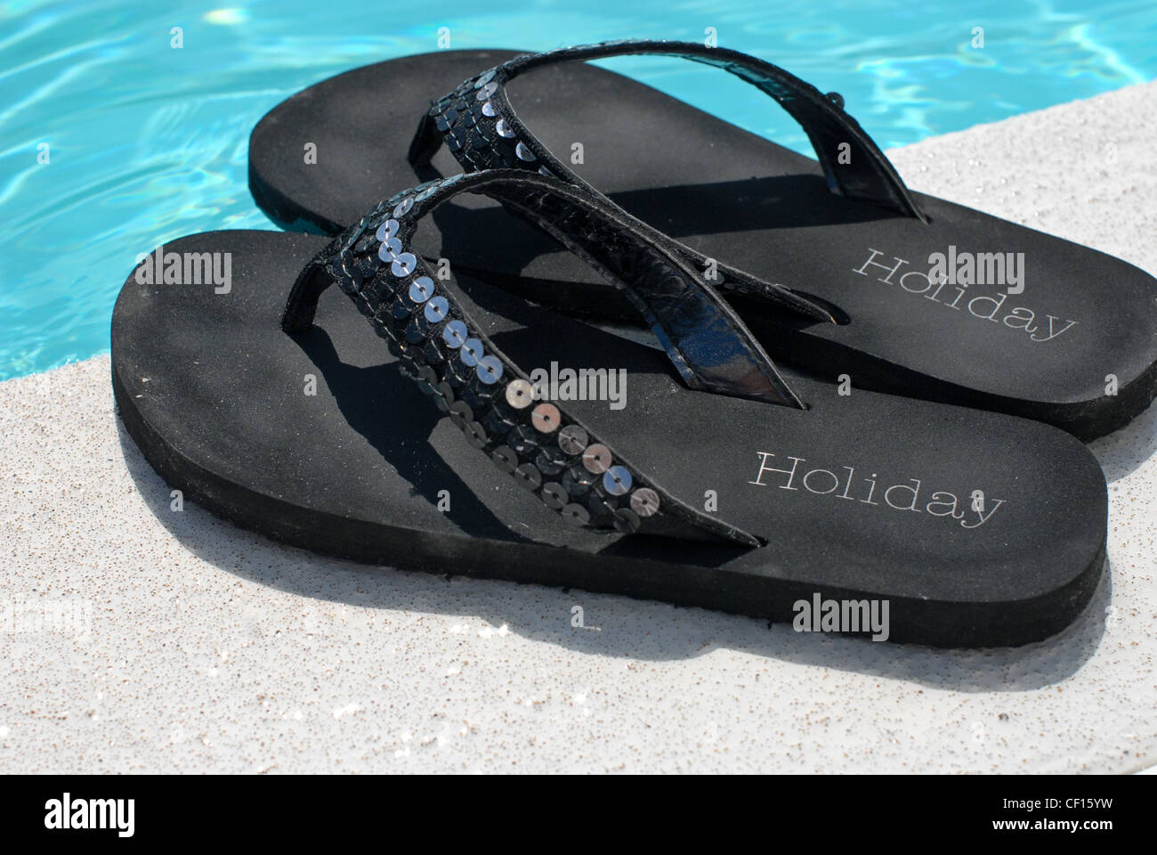 Ein paar schwarze Flip flops mit dem Wort Urlaub bedruckt links an der Seite eines Schwimmbades bei strahlendem Sonnenschein Stockfoto