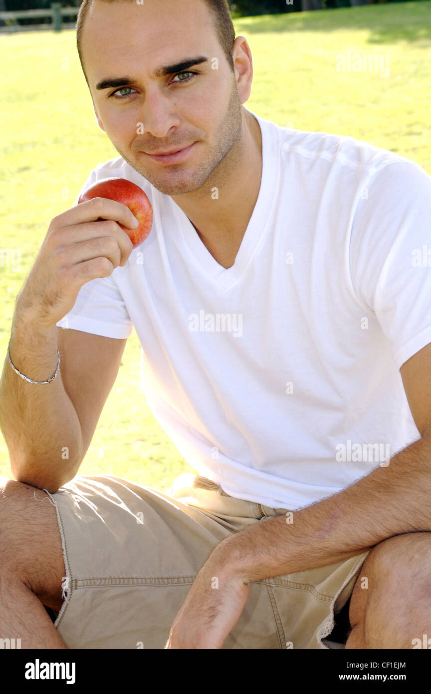 Männliche kurz getrimmten brünette Haare und leichte Stoppeln, ein weißes V-Hals-t-Shirt, Beige Shorts und Armband, einen Apfel essen Stockfoto