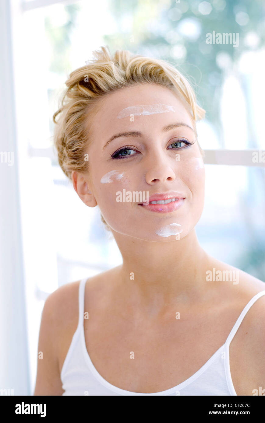 Weibliche blonde Haare tragen weiße Hemdchen Blobs Creme auf Stirn, Wangen und Kinn auf Kamera Lächeln zeigt Zähne Stockfoto