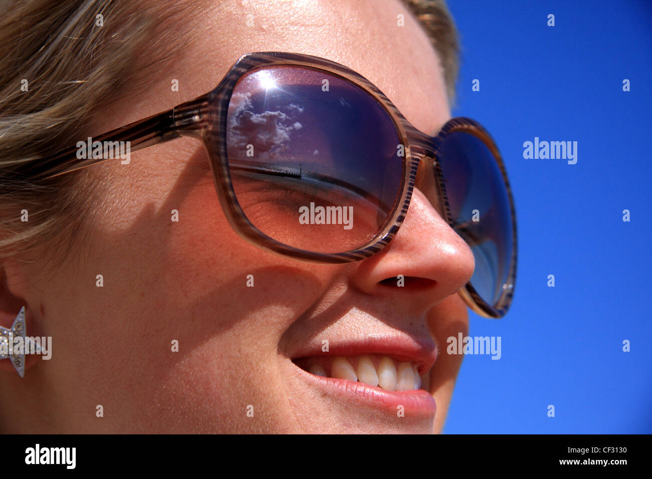 Weibchen tragen Sonnenbrillen und Sterne geformten Ohrring, Lächeln auf den Lippen gegen den blauen Himmel Stockfoto