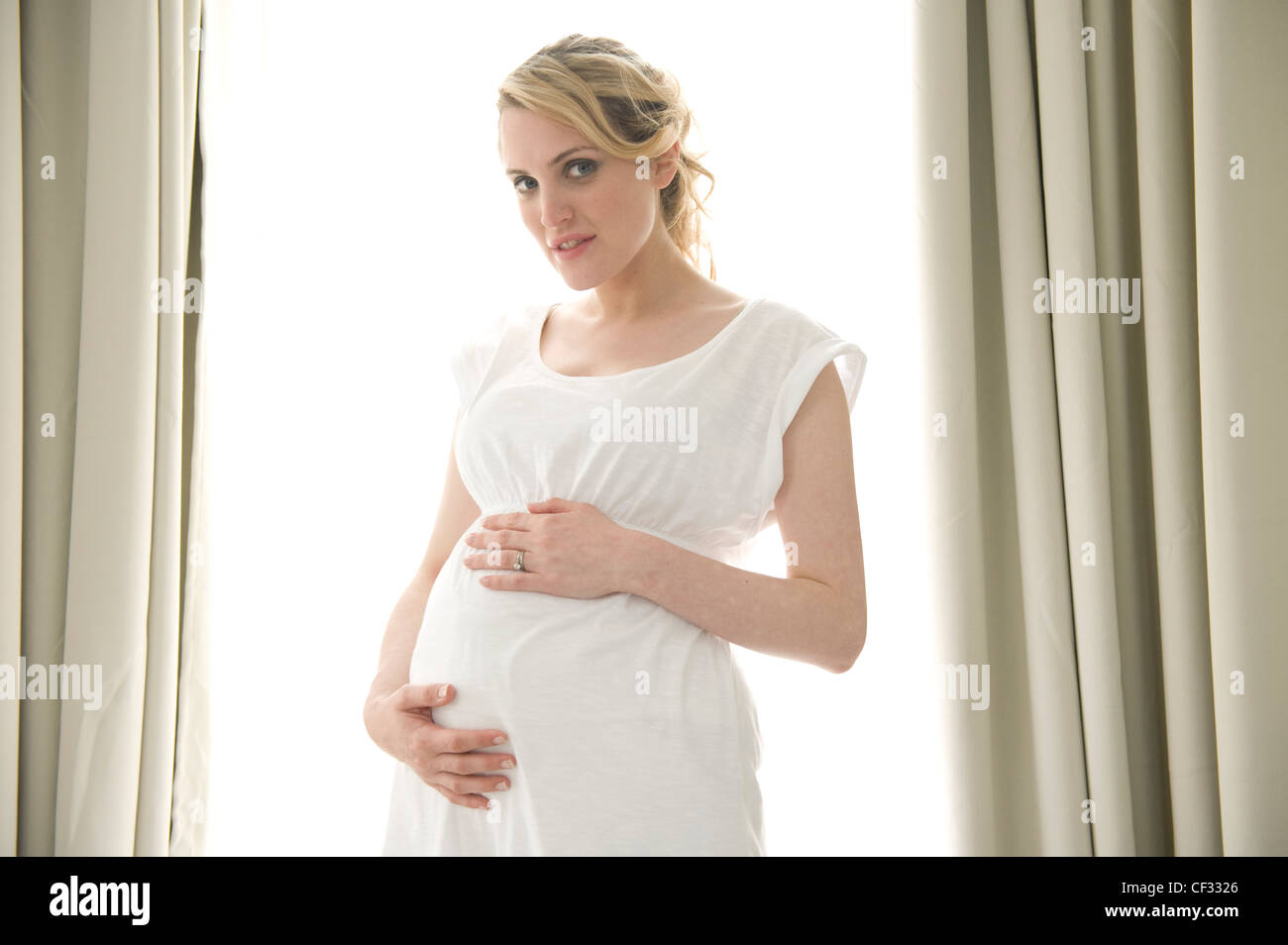 Schwangeres Frauen blondes Haar gebunden zu Pferdeschwanz, trägt ein weißes Kleid, stehend durch die Fenster Hände auf Beule, Lächeln, mit Blick auf Stockfoto