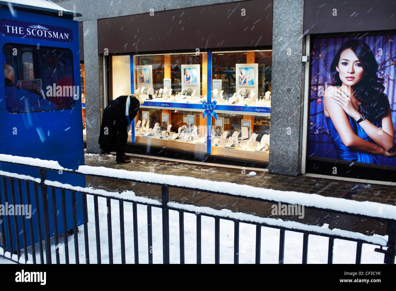 Eine Frau auf der Suche in der Auslage ein Juweliergeschäft gegenüber einen Zeitungskiosk, während der Schnee in der Princes Street fällt. Stockfoto