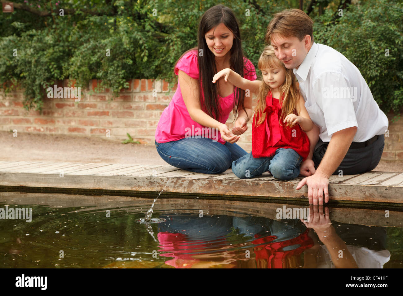 Kleines Mädchen im roten Kleid wirft Steinen im Wasser zusammen mit den Eltern. Stockfoto