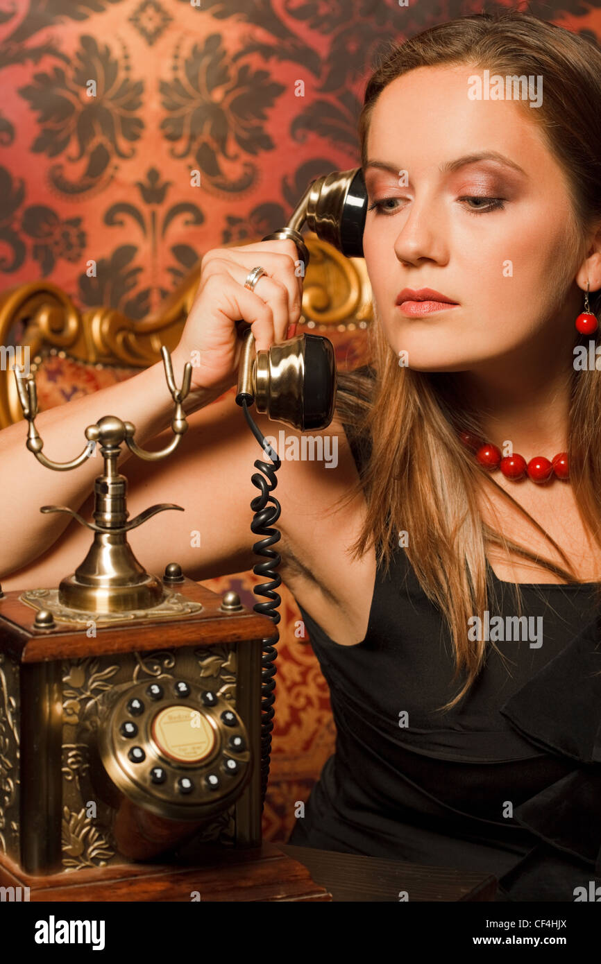 Frau im schwarzen Kleid auf einem Vintage Stuhl sitzen und am alten Telefon. das Rohr in seinen Händen hält und schaut Stockfoto