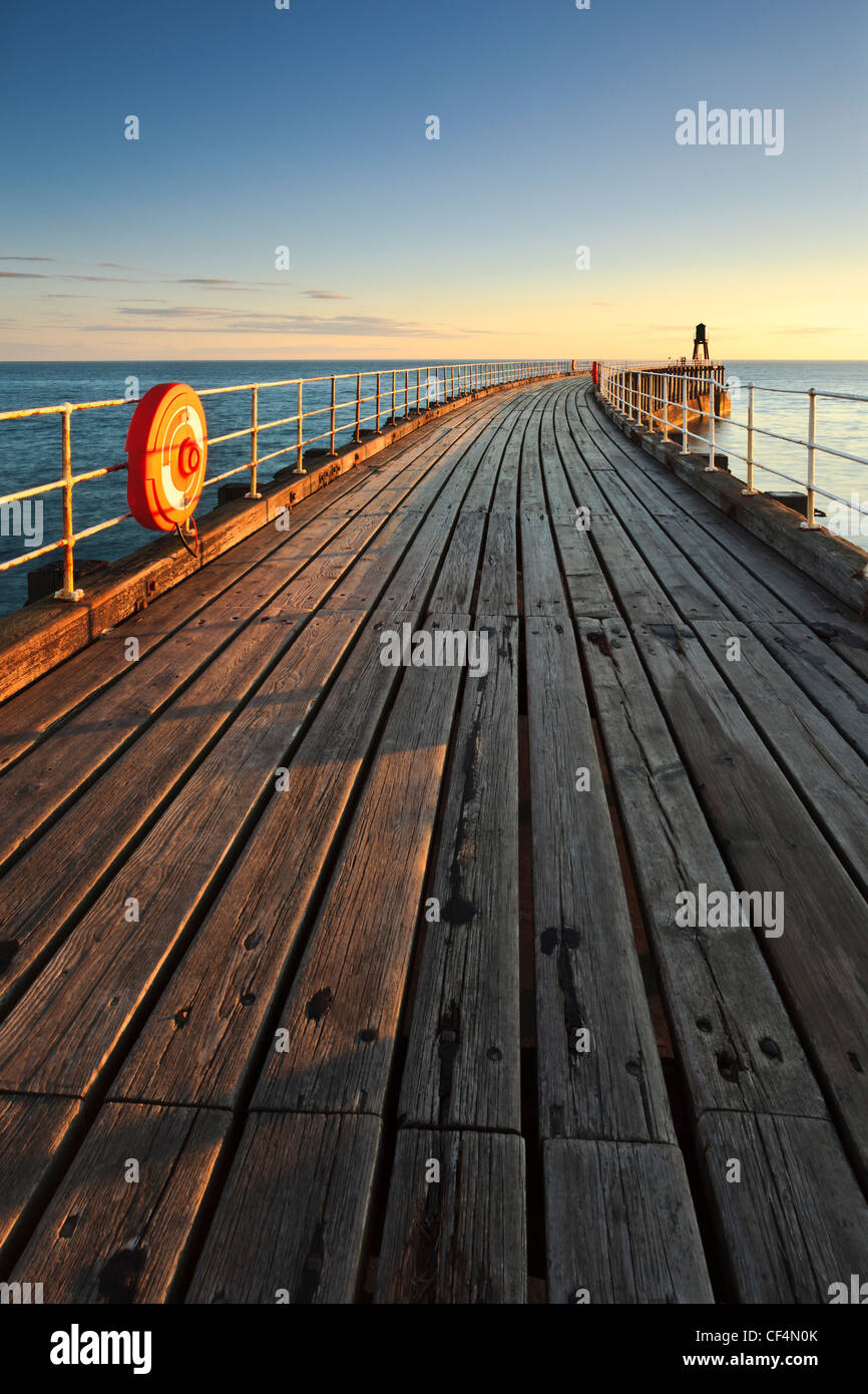 Zeigen Sie bei Sonnenaufgang entlang den Holzbrettern bis zum Leuchtturm am Ende der West Pier in Whitby an. Stockfoto