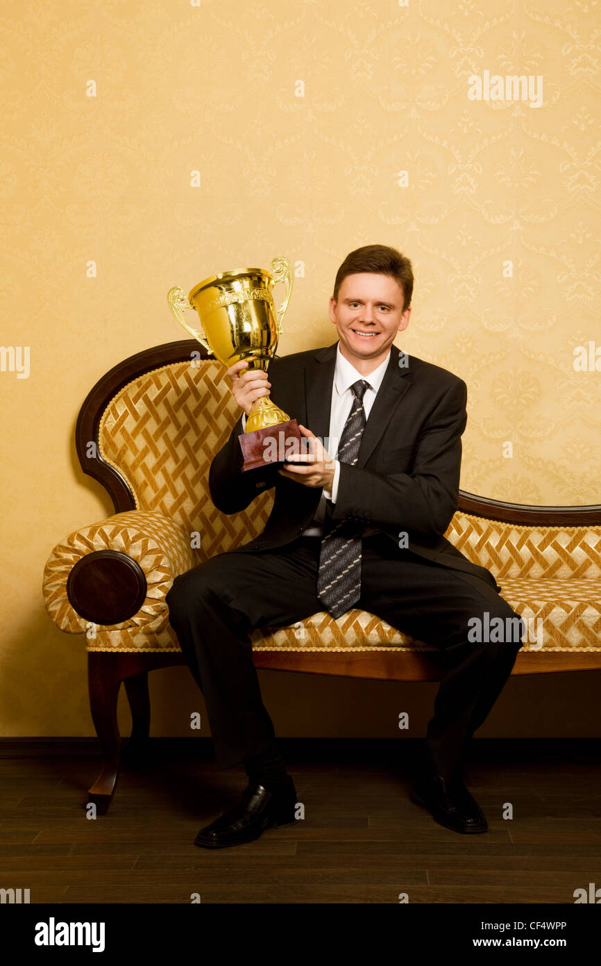 lächelnde Geschäftsmann mit Win-Cup in Hand im Anzug sitzt auf dem Sofa im Zimmer Stockfoto