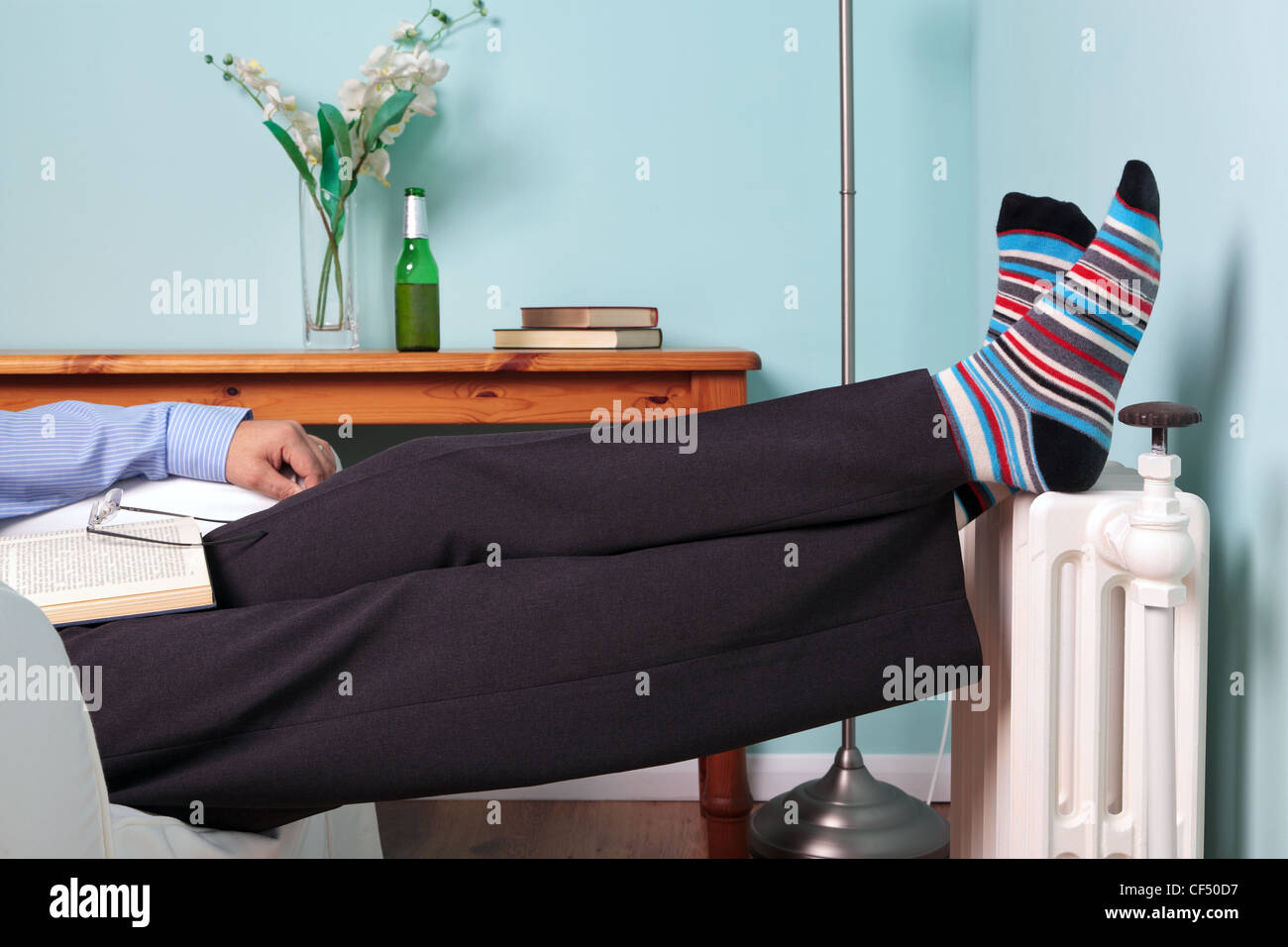Foto eines Mannes entspannend mit seinen Füßen oben auf einem Heizkörper mit einem Buch auf dem Schoß und Bier auf dem Tisch. Stockfoto